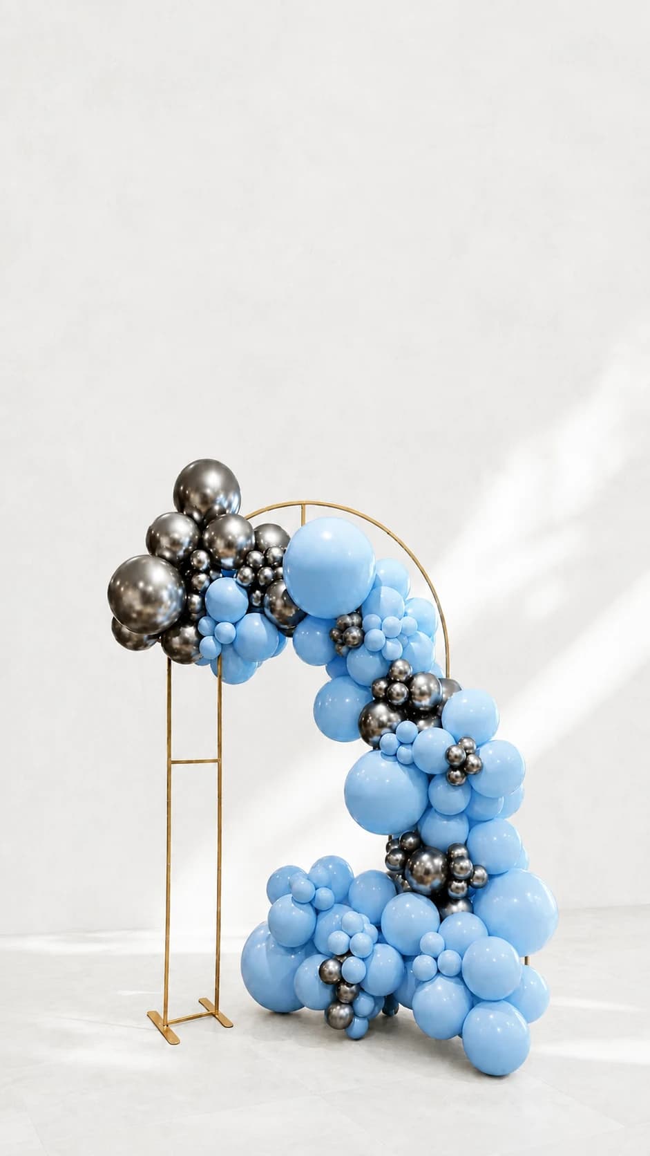 Blue, silver, and charcoal balloons on a gold circle stand, shown against an edited neutral background