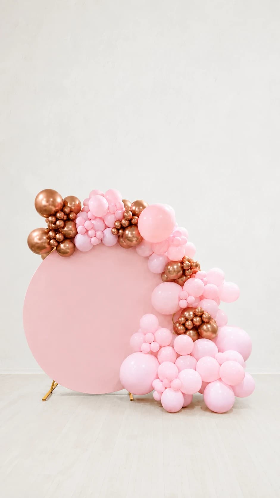 Pink and rose-gold balloon garland around a round blush backdrop, shown against an edited neutral background